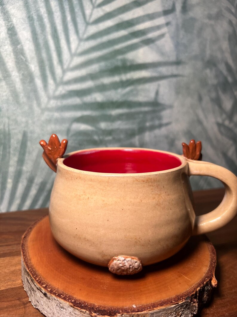 May include: A tan ceramic mug with a red interior, featuring small brown antler-like handles and a textured brown nose. The mug is placed on a round wooden coaster, perfect for coffee or tea.