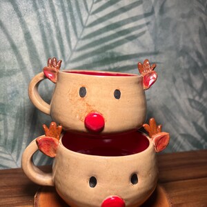 May include: Two stacked ceramic mugs with reindeer faces. Each mug has a tan exterior, red interior, black eyes, and a red nose. The mugs have brown antlers and handles. The mugs are on a wooden coaster.