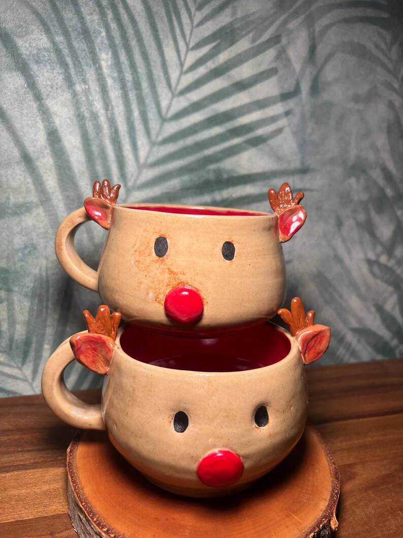 May include: Two stacked ceramic mugs with reindeer faces. Each mug has a tan exterior, red interior, black eyes, and a red nose. The mugs have brown antlers and handles. The mugs are on a wooden coaster.