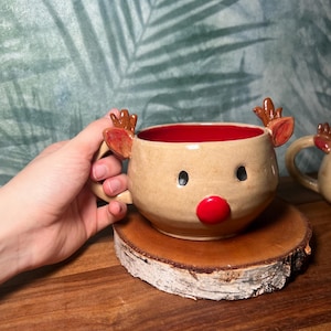 May include: A handmade ceramic mug shaped like a reindeer, with a tan exterior, red interior, and a red nose. The mug has small antlers and black eyes. It is sitting on a wooden coaster.