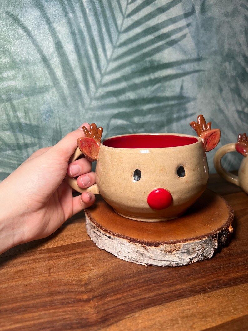 May include: A handmade ceramic mug shaped like a reindeer, with a tan exterior, red interior, and a red nose. The mug has small antlers and black eyes. It is sitting on a wooden coaster.