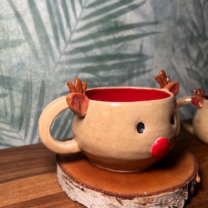 May include: A handmade ceramic reindeer mug with a tan exterior, red interior, and a round handle. The mug features a red nose, black eyes, and brown antlers. It sits on a small, round wooden coaster.