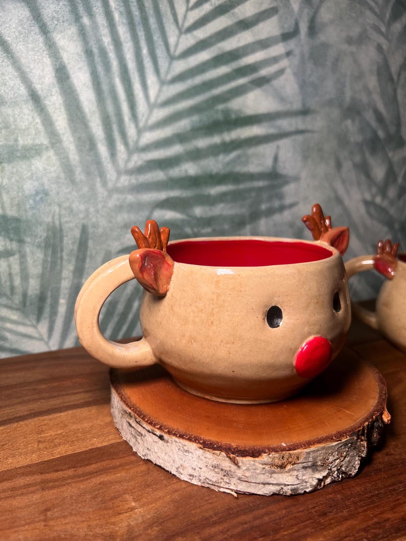 May include: A handmade ceramic reindeer mug with a tan exterior, red interior, and a round handle. The mug features a red nose, black eyes, and brown antlers. It sits on a small, round wooden coaster.
