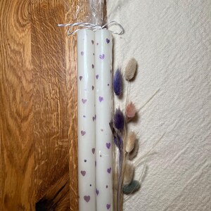 May include: Two white pillar candles decorated with purple hearts and dots, tied together with clear cellophane and twine. A small bundle of dried flowers in shades of purple, pink, and beige is included.