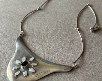 Puede incluir: Un collar plateado con un colgante asimétrico único. El colgante presenta una piedra negra central rodeada de piedras opalescentes en forma de flor. El collar tiene una cadena fina y un cierre.