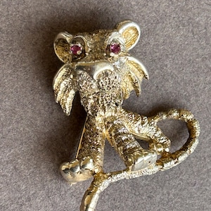 Op de afbeelding: Een goudkleurige metalen broche in de vorm van een gestileerde leeuw. De leeuw heeft rode edelsteenogen en gedetailleerde texturen. De broche is ontworpen met een speld aan de achterkant voor een veilige bevestiging.