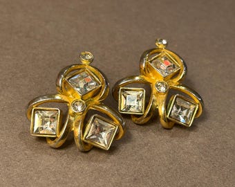 Op de afbeelding: Een paar gouden oorbellen met heldere kristallen. De oorbellen zijn gevormd als een klavertjevier met een kristal in het midden van elk blad.