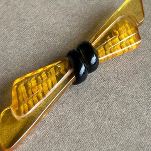 Könnte beinhalten: Bernsteinfarbene Vintage-Brosche in Schleifenform. Die Brosche hat ein strukturiertes, geripptes Muster und wird von zwei schwarzen, glänzenden Ringen in der Mitte zusammengehalten. Ein einzigartiges Accessoire.