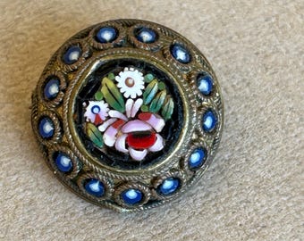 Puede incluir: Broche redondo vintage con diseño floral de mosaico. El centro presenta una disposición detallada de flores rosas, rojas, blancas y verdes sobre un fondo negro. El broche está enmarcado por un borde de metal dorado con detalles azules.