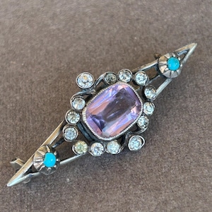 May include: A vintage silver brooch with a central rectangular amethyst stone, surrounded by clear crystals and turquoise accents. The brooch has a symmetrical design with pointed ends, showcasing a classic, elegant style.