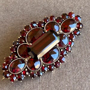 May include: A large, antique brooch with a central, rectangular, dark red gemstone surrounded by smaller, round, dark red gemstones. The brooch is set in silver.