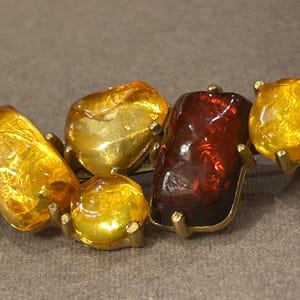 Puede incluir: Broche vintage con cinco piedras de color ámbar de formas irregulares. Las piedras están engastadas en una base de metal dorado. El broche tiene una paleta de colores cálidos, con tonos de amarillo, naranja y rojo.