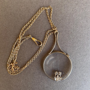 Puede incluir: Una lupa colgante vintage con una cadena dorada y un marco plateado. La lupa redonda tiene un diseño floral en la parte inferior. La cadena tiene un cierre.