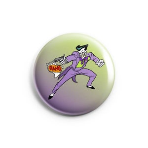 May include: A purple and green button with a cartoon image of the Joker from Batman. The Joker is holding a gun and a sign that says 'BANG' in red letters.