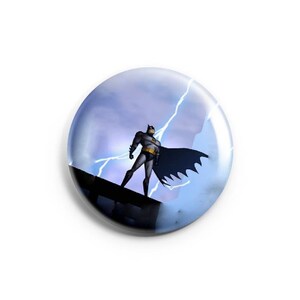 May include: A round button with a blue and white background featuring a cartoon image of Batman standing on a rooftop with lightning bolts in the background.