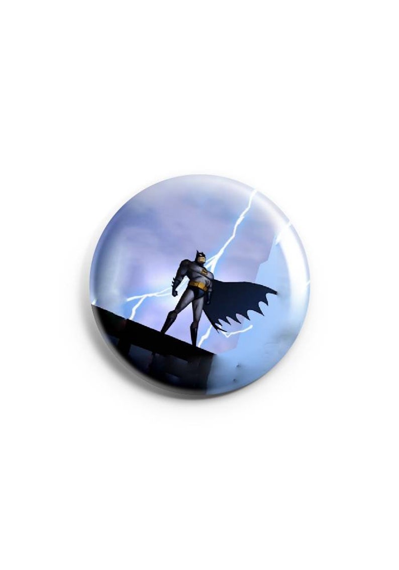 May include: A round button with a blue and white background featuring a cartoon image of Batman standing on a rooftop with lightning bolts in the background.