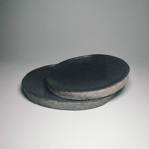 May include: Two black ceramic dishes, one stacked on top of the other. The top dish is slightly larger and has a rough, textured rim.