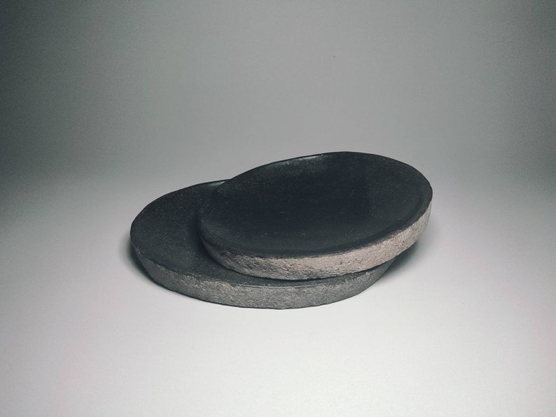 May include: Two black ceramic dishes, one stacked on top of the other. The top dish is slightly larger and has a rough, textured rim.