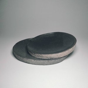 May include: Two black ceramic plates, one stacked on top of the other. The plates have a rough, textured surface and a slightly oval shape.