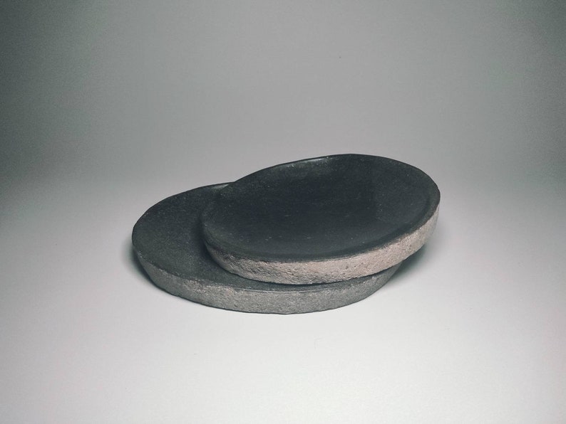 May include: Two black ceramic plates, one stacked on top of the other. The plates have a rough, textured surface and a slightly oval shape.