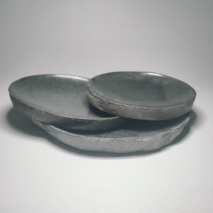 May include: Three grey ceramic dishes of varying sizes stacked on top of each other. The dishes have a rough, textured surface.