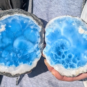 May include: Two halves of a polished gemstone, showcasing a vibrant blue interior with white and gray edges. The stone's surface has a swirling pattern, resembling ocean waves. The overall shape is round, with a rough, natural exterior.
