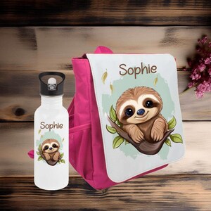 Könnte beinhalten: Eine weiße Wasserflasche und ein pinkfarbener Rucksack mit einem Faultiermotiv. Die Wasserflasche und der Rucksack tragen beide den Namen "Sophie" und eine Cartoon-Faultier-Illustration. Der Rucksack hat eine weiße Klappe mit dem Faultiermotiv.