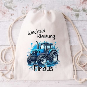 Könnte beinhalten: Eine weiße Kordeltasche mit dem Aufdruck "Wechsel Kleidung Findus". Die Tasche zeigt einen blauen Comic-Traktor mit Wasserspritzern drumherum.