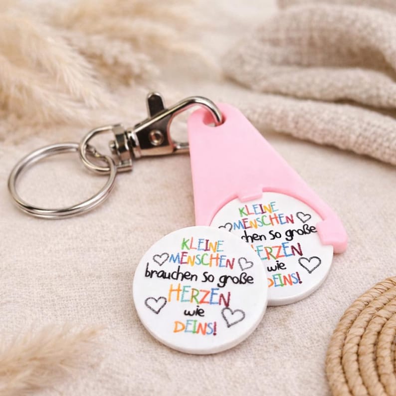 Puede incluir: Un llavero con un soporte rosa para monedas de carrito y dos fichas redondas blancas. Las fichas tienen el texto "KLEINE MENSCHEN brauchen so gro&szlig;e HERZEN wie DEINS!" en letras coloridas e ilustraciones de corazones. El llavero tiene un mosquet&oacute;n y un anillo plateados.