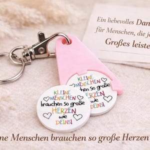 Puede incluir: Un llavero con un soporte rosa para fichas de carrito de la compra y dos fichas blancas. Las fichas tienen texto colorido e ilustraciones de corazones. El texto de las fichas dice "Kleine Menschen brauchen so gro&szlig;e Herzen wie deins!"