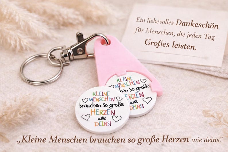 Puede incluir: Un llavero con un soporte rosa para fichas de carrito de la compra y dos fichas blancas. Las fichas tienen texto colorido e ilustraciones de corazones. El texto de las fichas dice "Kleine Menschen brauchen so gro&szlig;e Herzen wie deins!"