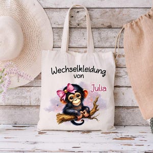 Könnte beinhalten: Eine weiße Stofftasche mit dem Text "Wechselkleidung von Julia" und einem Cartoon-Bild eines Affen mit einer rosa Schleife.