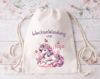 Könnte beinhalten: Ein weißer Kordelzugbeutel mit einem lila Einhorn und Schmetterlingen darauf gedruckt. Die Tasche sagt "Wechselkleidung von Raja".