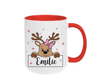Könnte beinhalten: Weiße Keramiktasse mit rotem Rand und Henkel. Die Tasse zeigt ein Cartoon-Rentier mit einer rosa Schleife, das über ein Schild mit der Aufschrift "Emilie" schaut.