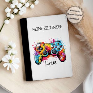 Op de afbeelding: Wit notitieboek met de tekst "MEINE ZEUGNISSE" en een kleurrijk videogamecontroller-ontwerp. De controller is versierd met verfspetters en de naam "Linus" staat eronder. Het notitieboek heeft een zwarte rug.