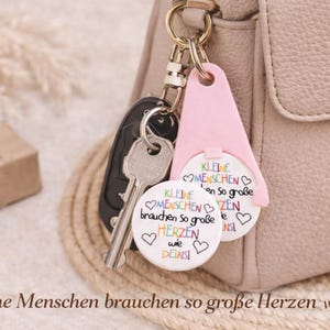 Puede incluir: Un llavero con una etiqueta rosa y dos colgantes circulares blancos, sujeto a una llave de coche y a un bolso. Los colgantes tienen el texto "Kleine Menschen brauchen so gro&szlig;e Herzen wie deins" y corazones coloridos.