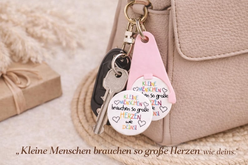 Puede incluir: Un llavero con una etiqueta rosa y dos colgantes circulares blancos, sujeto a una llave de coche y a un bolso. Los colgantes tienen el texto "Kleine Menschen brauchen so gro&szlig;e Herzen wie deins" y corazones coloridos.