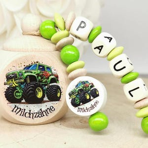 Op de afbeelding: Een houten doos met een groen monstertruck-ontwerp en de tekst "Milchzähne" op de voorkant. Een houten kralenketting met groene en witte kralen en de naam "PAUL" gespeld op de kralen.