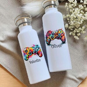 Könnte beinhalten: Zwei weiße Wasserflaschen mit Bambusdeckeln. Jede Flasche hat ein farbenfrohes Aquarelldesign eines Videospiel-Controllers. Auf einer Flasche steht der Name "Valentin", auf der anderen "Oliver".