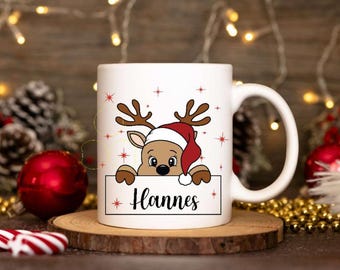 Könnte beinhalten: Weiße Keramiktasse mit einem Rentier-Motiv. Das Rentier trägt einen roten Weihnachtsmann-Hut und schaut über ein weißes Schild, auf dem "Hannes" steht.