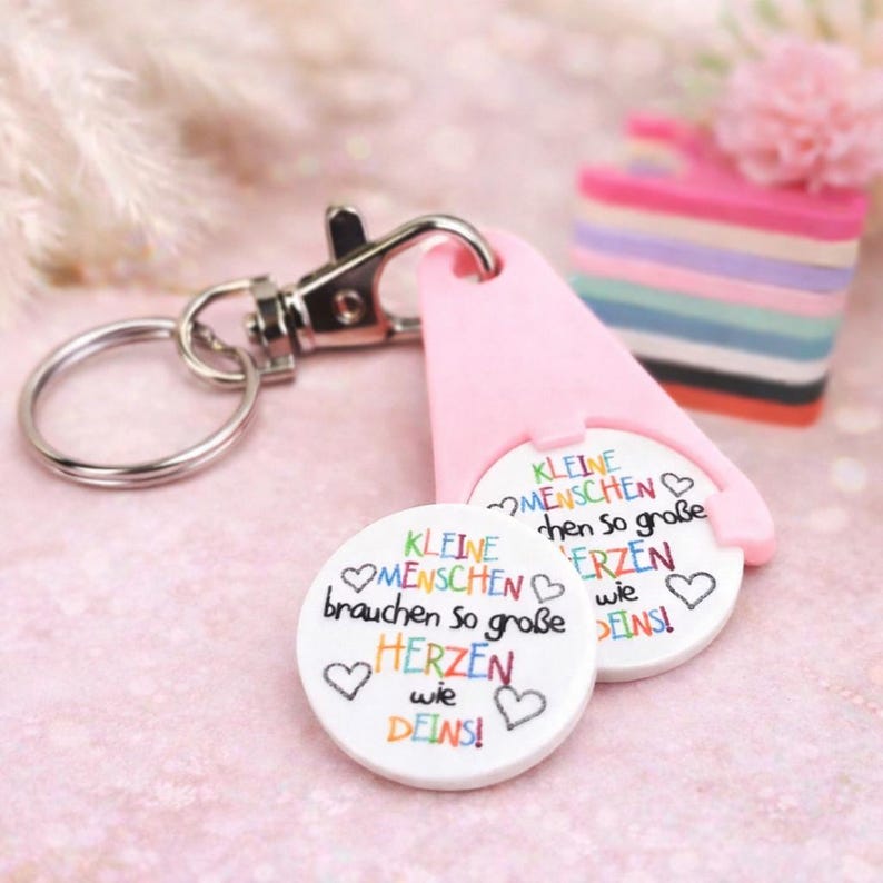 Puede incluir: Un llavero rosa y plateado con dos fichas circulares blancas. Las fichas tienen el texto alem&aacute;n "KLEINE MENSCHEN brauchen so gro&szlig;e HERZEN wie DEINS!" en letras coloridas. Un soporte de ficha rosa en forma de tri&aacute;ngulo est&aacute; adjunto.