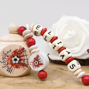 Op de afbeelding: Een lichtgekleurde houten houder met deksel, voorzien van een voetbalafbeelding en de Duitse tekst "Milchzähne". Er is een snoer van rode, witte en naturel houten kralen aan bevestigd die de naam "ELIAAS" vormen met zwarte letters.