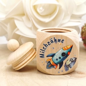 Op de afbeelding: Een houten doos met een deksel, versierd met een raket en sterren. De doos is gelabeld met "Milchzähne", wat Duits is voor "melkgebit".