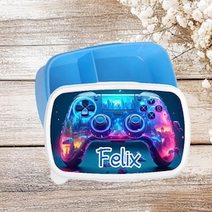 Op de afbeelding: Een blauwe lunchbox met een wit deksel met een kleurrijke afbeelding van een videogamecontroller met de naam "Felix" eronder gedrukt.