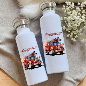 Könnte beinhalten: Zwei weiße Wasserflaschen mit silbernen Verschlüssen und Bambusdeckeln. Jede Flasche hat eine rote Feuerwehrauto-Grafik und den Namen "Aleksander" in roter Schrift. Die Flaschen stehen auf einer grauen Oberfläche.