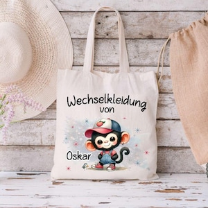 Könnte beinhalten: Eine weiße Stofftasche mit dem Text "Wechselkleidung von Oskar" und einem Comic-Affen, der ein rot-blaues Outfit und eine Baseballkappe trägt.