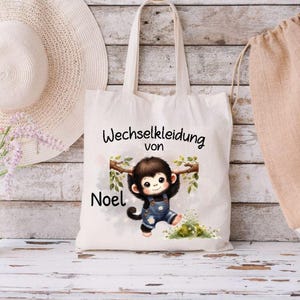 Könnte beinhalten: Eine cremefarbene Tragetasche mit einem Cartoon-Affen-Design. Der Affe trägt eine blaue Latzhose und hängt an einem Ast. Der Text "Wechselkleidung von Noel" ist über dem Affen aufgedruckt.