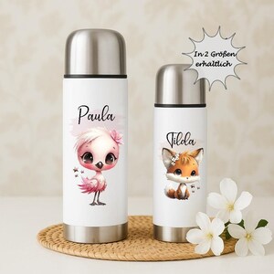 Könnte beinhalten: Zwei weiße isolierte Wasserflaschen mit silbernen Deckeln. Eine zeigt eine Flamingo-Illustration und den Namen "Paula", die andere eine Fuchs-Illustration und den Namen "Tilda". Beide Flaschen stehen auf einer gewebten Matte.