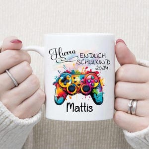 Könnte beinhalten: Weiße Keramiktasse mit einem farbenfrohen Aquarelldesign eines Videospielcontrollers. Der Text "Hurra Endlich Schulkind 2024 Mattis" ist auf die Tasse gedruckt.