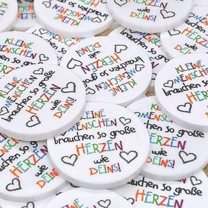 Puede incluir: Una colecci&oacute;n de objetos circulares blancos con la frase alemana "Kleine Menschen brauchen so gro&szlig;e Herzen wie Deins!" en varios colores. Cada disco tiene un s&iacute;mbolo de coraz&oacute;n.